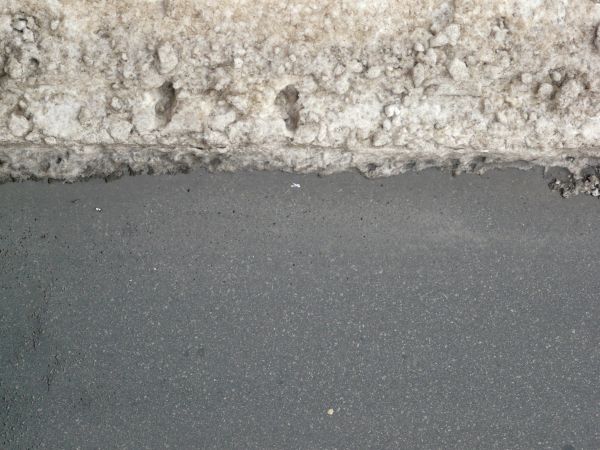 Road edge texture, grey in color with tiny rocks embedded within it, meeting an uneven area of clumped white and brown snow.